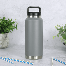 Load image into Gallery viewer, grey stainless steel water bottle on concrete surface with grey and blue striped drinking paper straws in the foreground with foliage in the background
