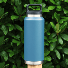 Load image into Gallery viewer, blue large stainless steel water bottle with carry handle lid on fence post outdoors in front of leafy foliage
