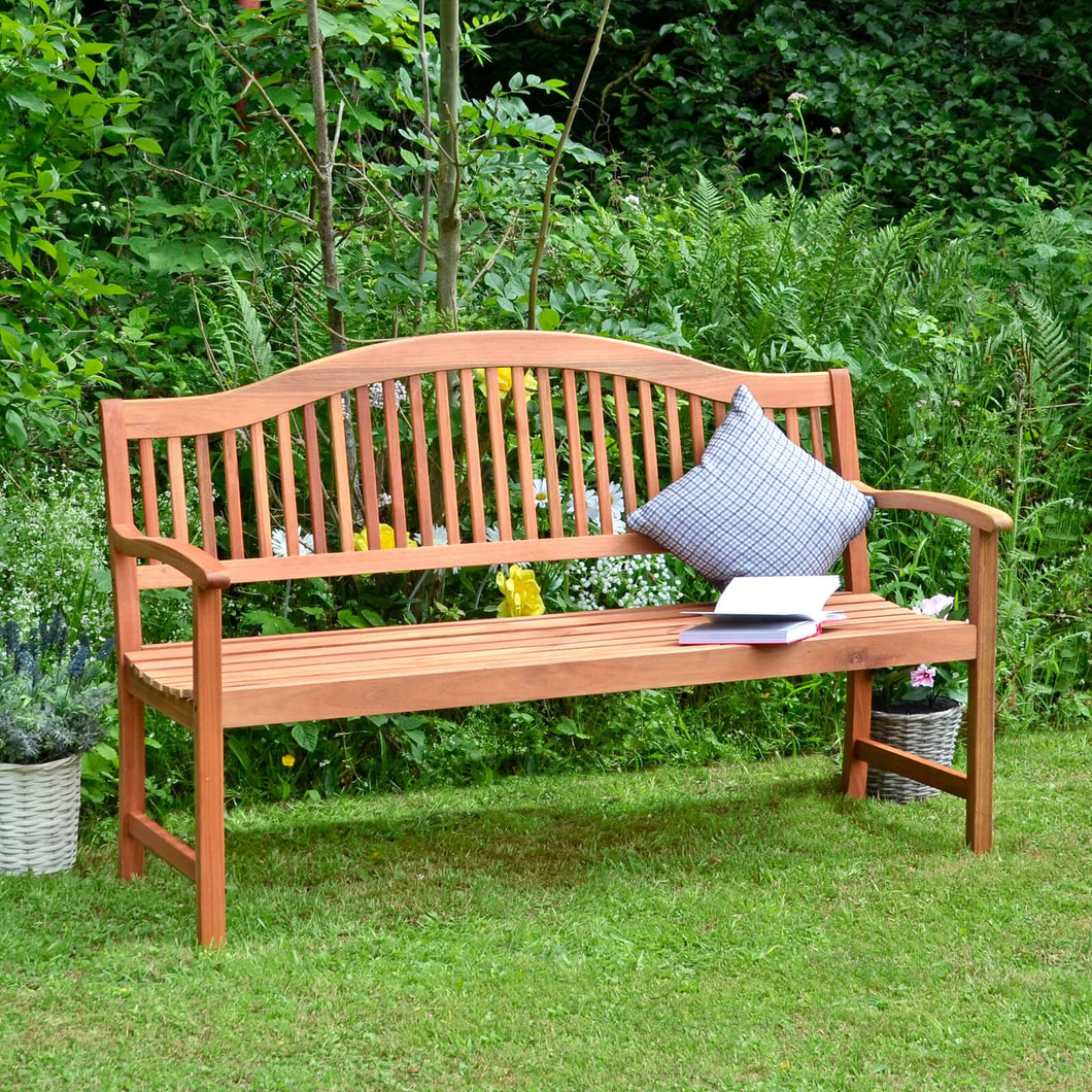 Large 3 seater wooden garden bench, traditional style with slatted back sitting on grass in a garden with cushion, book, flowers and plants