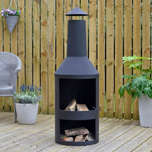 Load image into Gallery viewer, Black chiminea style log burner with log storage, on garden decking with plants, grey armchair and logs ready for burning
