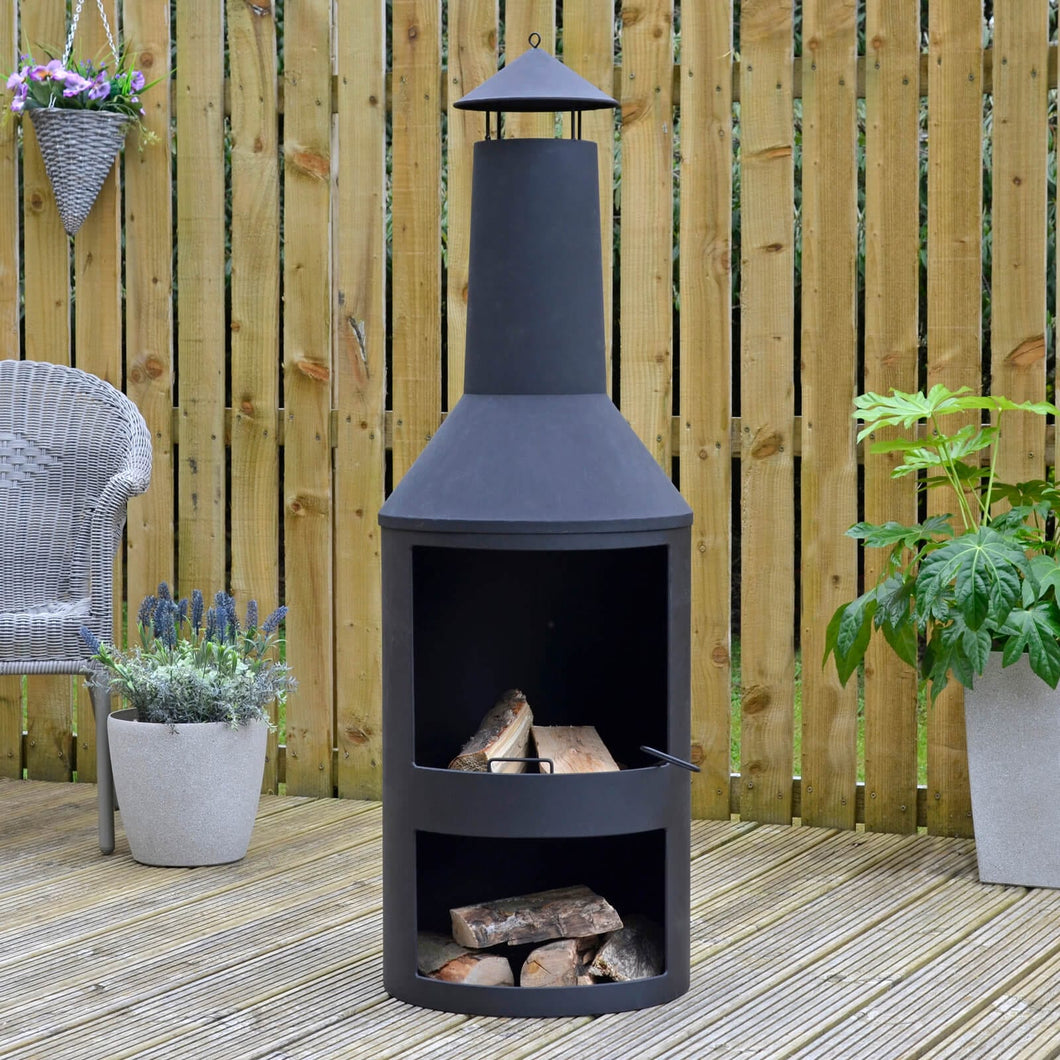 Black chiminea style log burner with log storage, on garden decking with plants, grey armchair and logs ready for burning