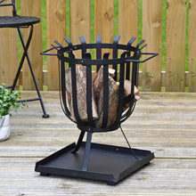 Load image into Gallery viewer, Brazier style wood burning fire pit with 3 legs and open grate with ash collection tray, filled with logs ready for burning, sitting on decking with plants and black metal bistro chair
