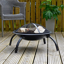 Load image into Gallery viewer, Black Vulcan fire pit with fold out legs and mesh spark cover, filled with wood logs ready for burning, sitting on decking with grey rattan garden chair and plants
