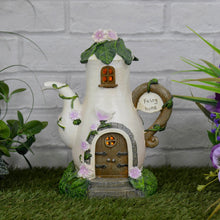 Load image into Gallery viewer, Solar coffee pot garden ornament lit up by orange light on grass beside white brick wall and flowers
