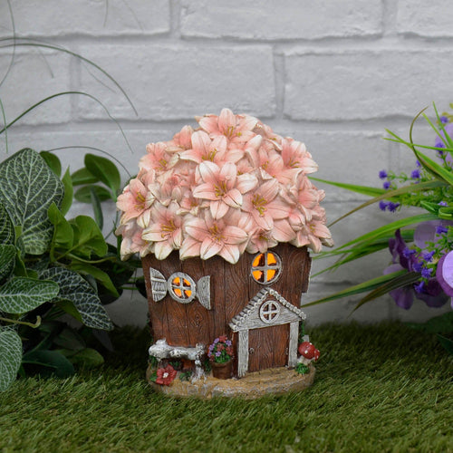 Solar cottage lit up by orange light inside, with pink blossom roof sitting on grass beside a white brick wall and green plants