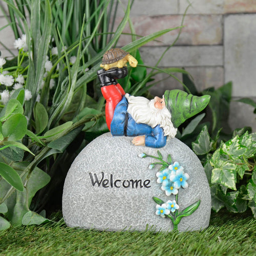 Garden gnome ornament, with grey stone pebble, Welcome text, blue flowers, figure with green hat and tortoise on his boots, in a garden with grass an dplants