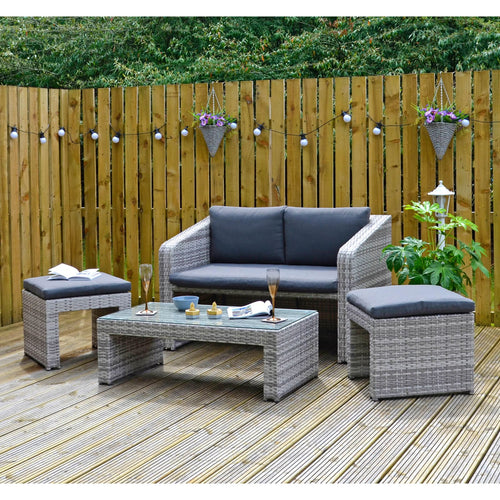 Grey rattan garden sofa set with 2 seater sofa, 2 ottoman stools and a glass topped coffee table, on decking with wine glasses magazine, plants and party lights