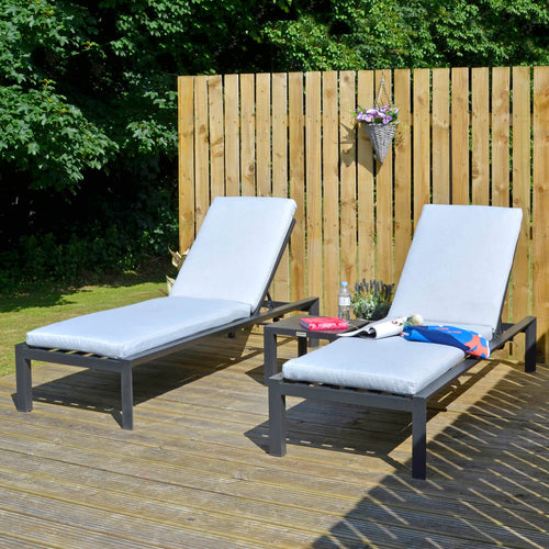 Set of 2 sun loungers and table with dark grey frames and light grey cushions, on garden decking with towel, water and plants